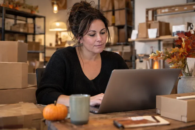 A shop owner works at a laptop among boxes in a stockroom.